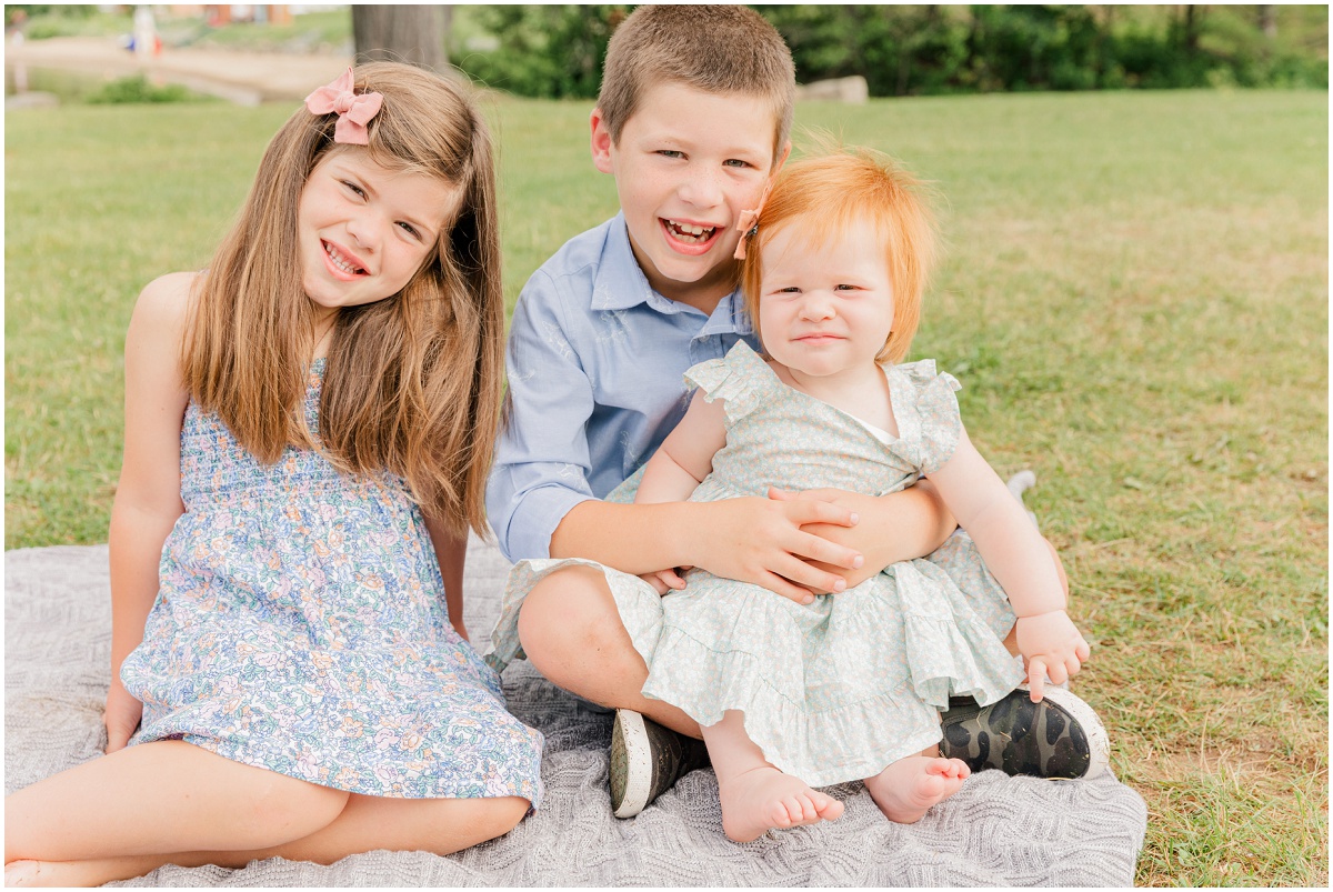 Three children sit on a blanket and smile at the camera