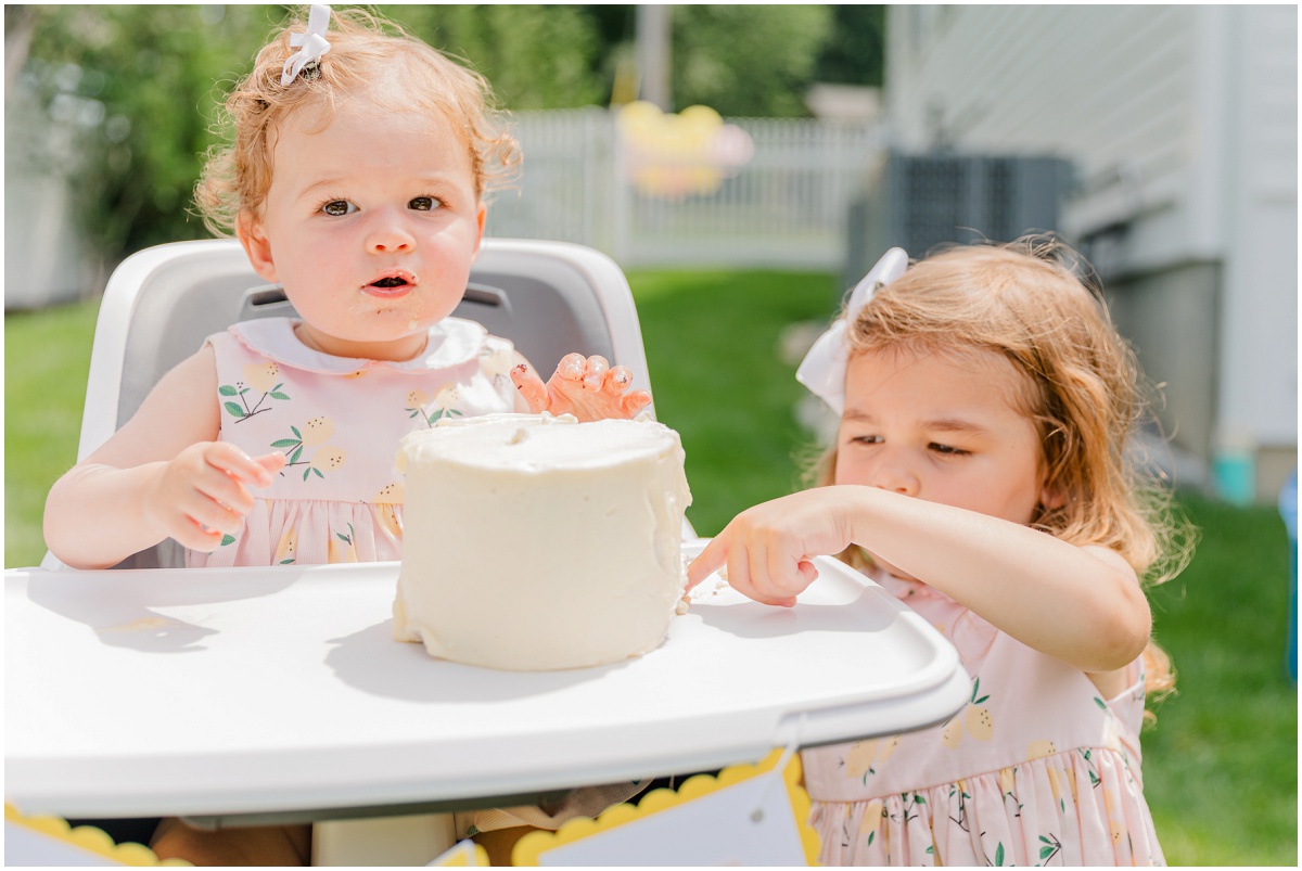 One-year-old smiling in high chair and sister tasting smash cake frosting