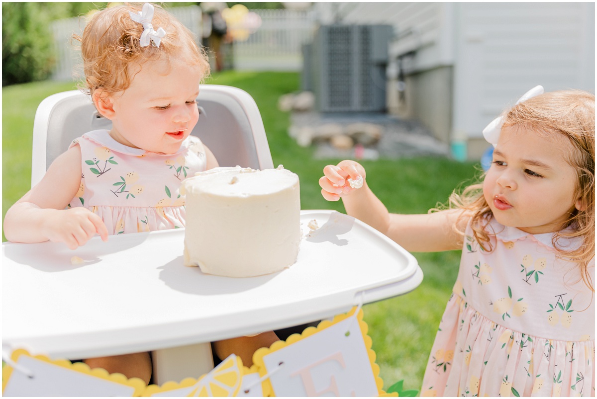 One-year-old smiling in high chair and sister tasting smash cake frosting