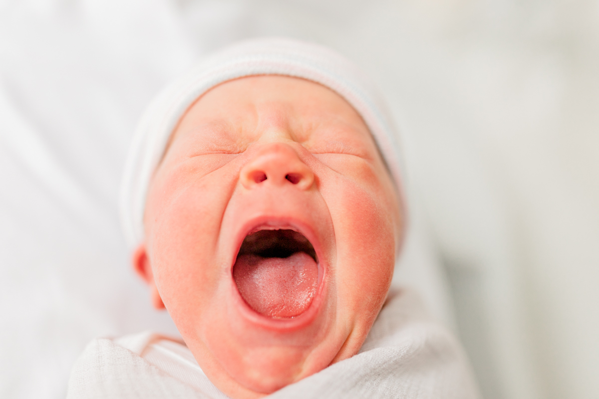 Close-up of newborn baby yawning; baby is wearing a hospital hat and is in a light grey swaddle in the hospital bassinet