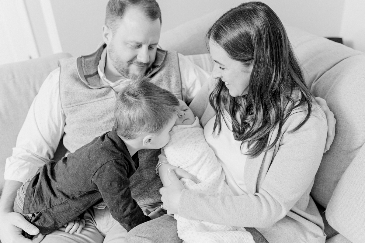 Home Birth in Massachusetts - home environment comforts | black and white image of a family of 4 sitting on a couch. Mother and father sitting next to each other, smiling down at newborn in Mom's arms while toddler big brother leans in to kiss the newborn.