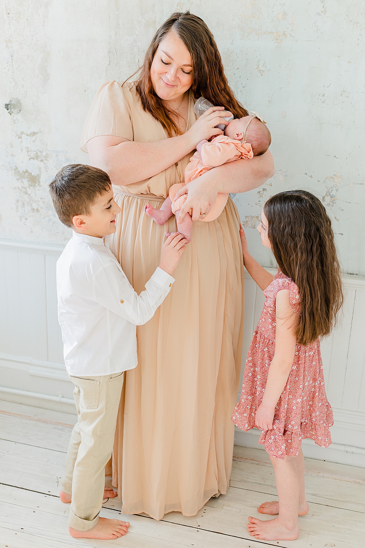Newborn Photography in the North Shore | Mother in long neutral dress standing and feeding a newborn baby a bottle while brother tickles baby's feet and sister looks on