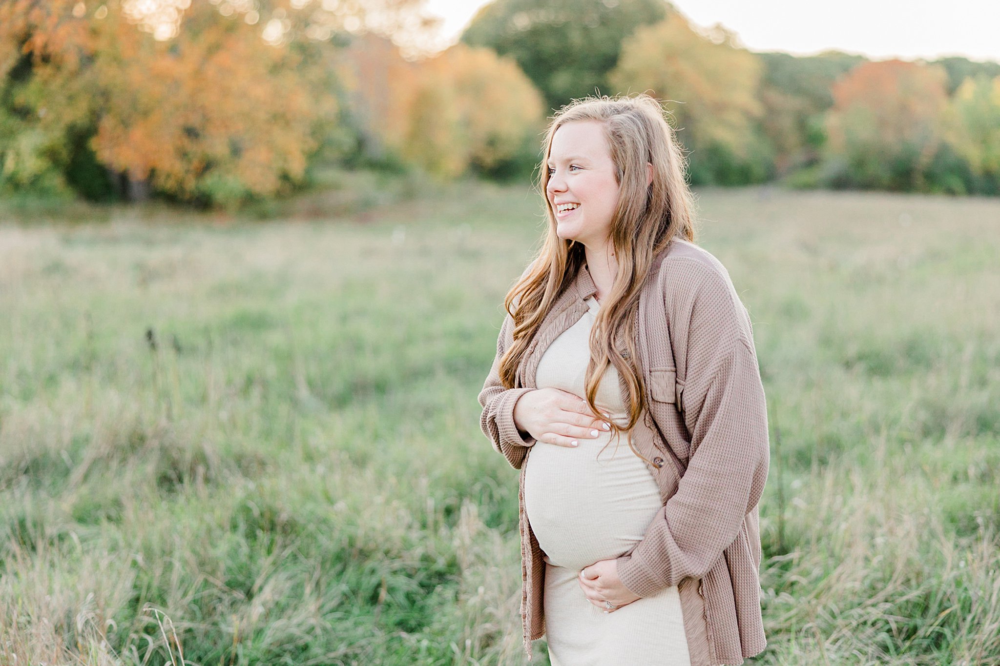 A mother to be in a beige dress and tan sweater holds her bump while standing in a green grassy field prenatal chiropractor