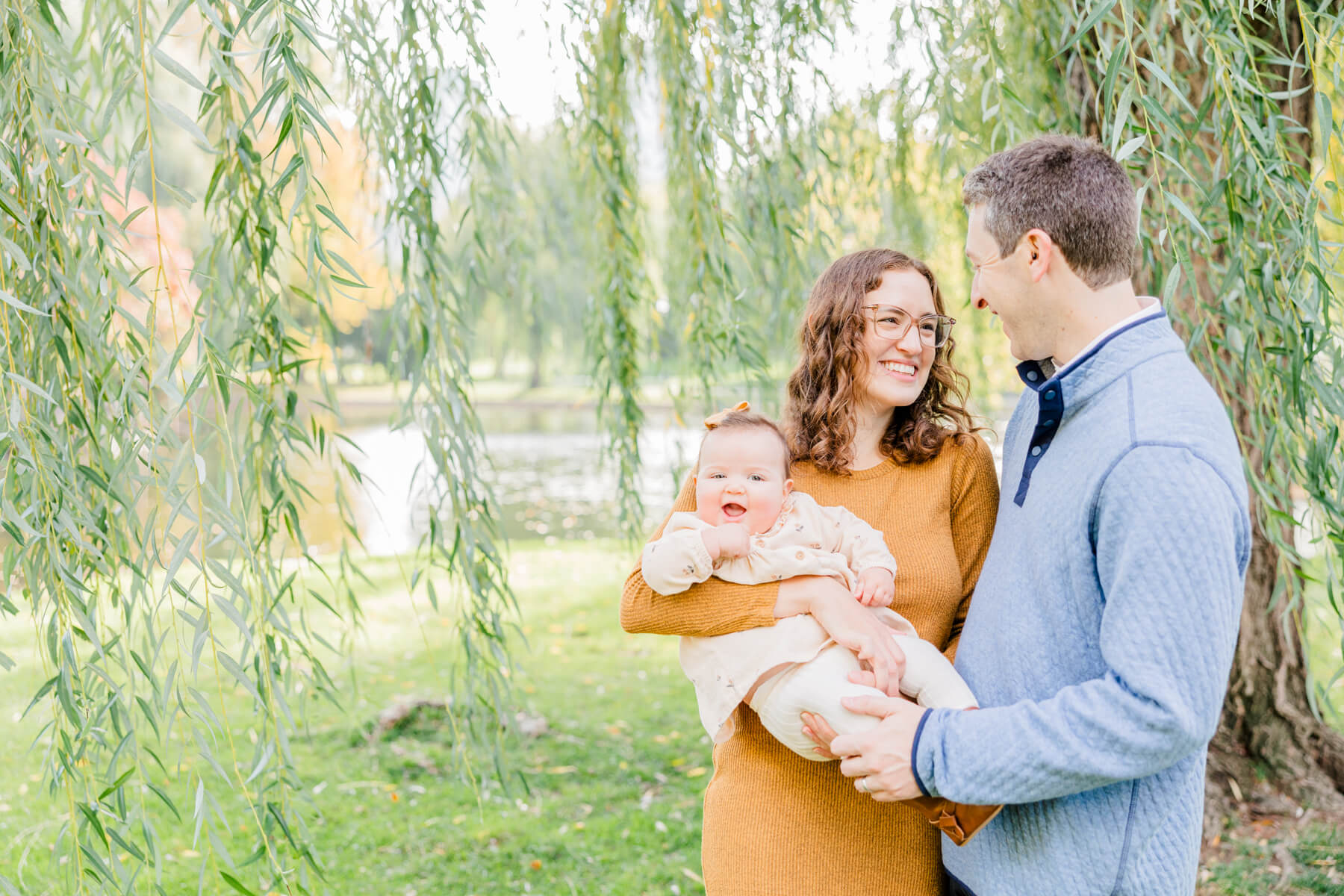New parents stand under a willow tree by a lake holding their smiling toddler girl brigham fertility clinic
