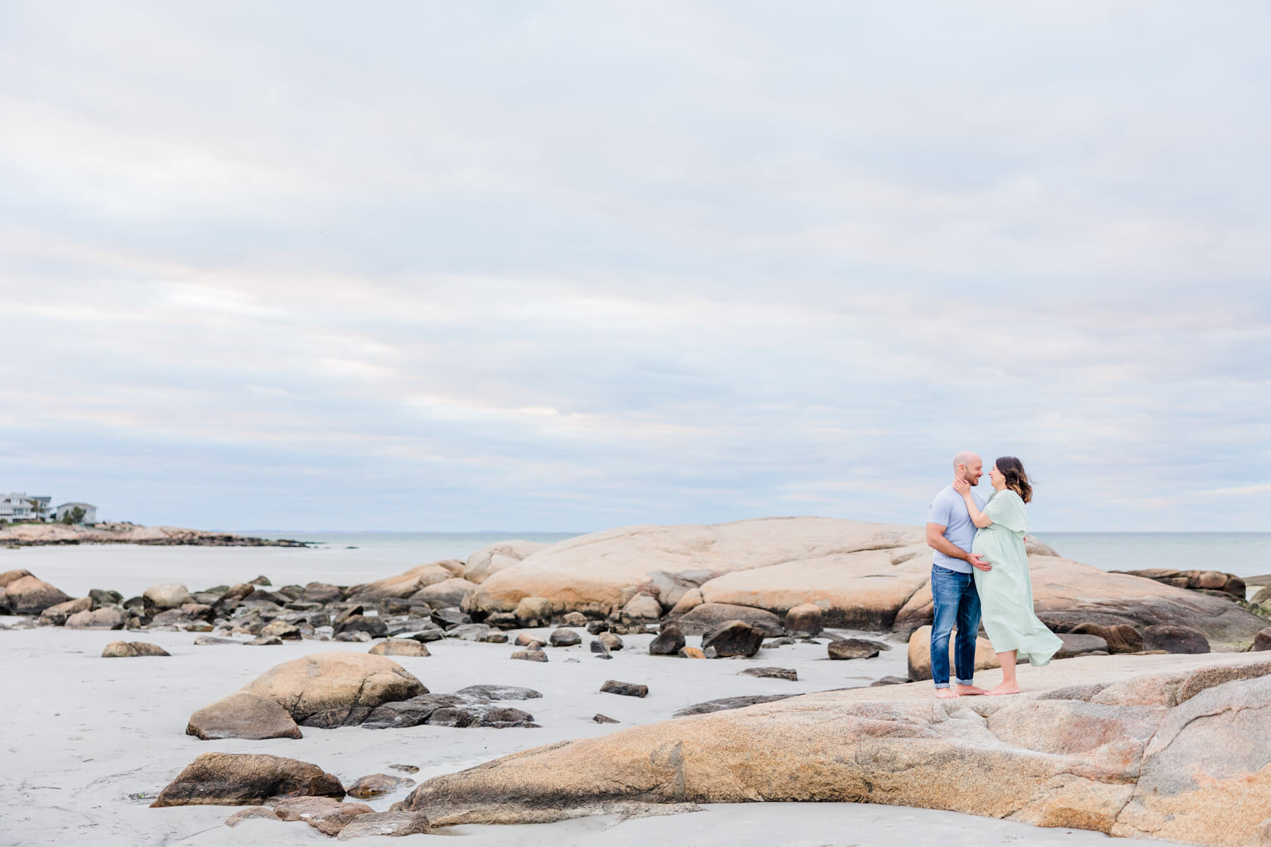 A mother and father stand on a rock in the water while holding the bump and looking into each other's eyes obgyn leominster ma