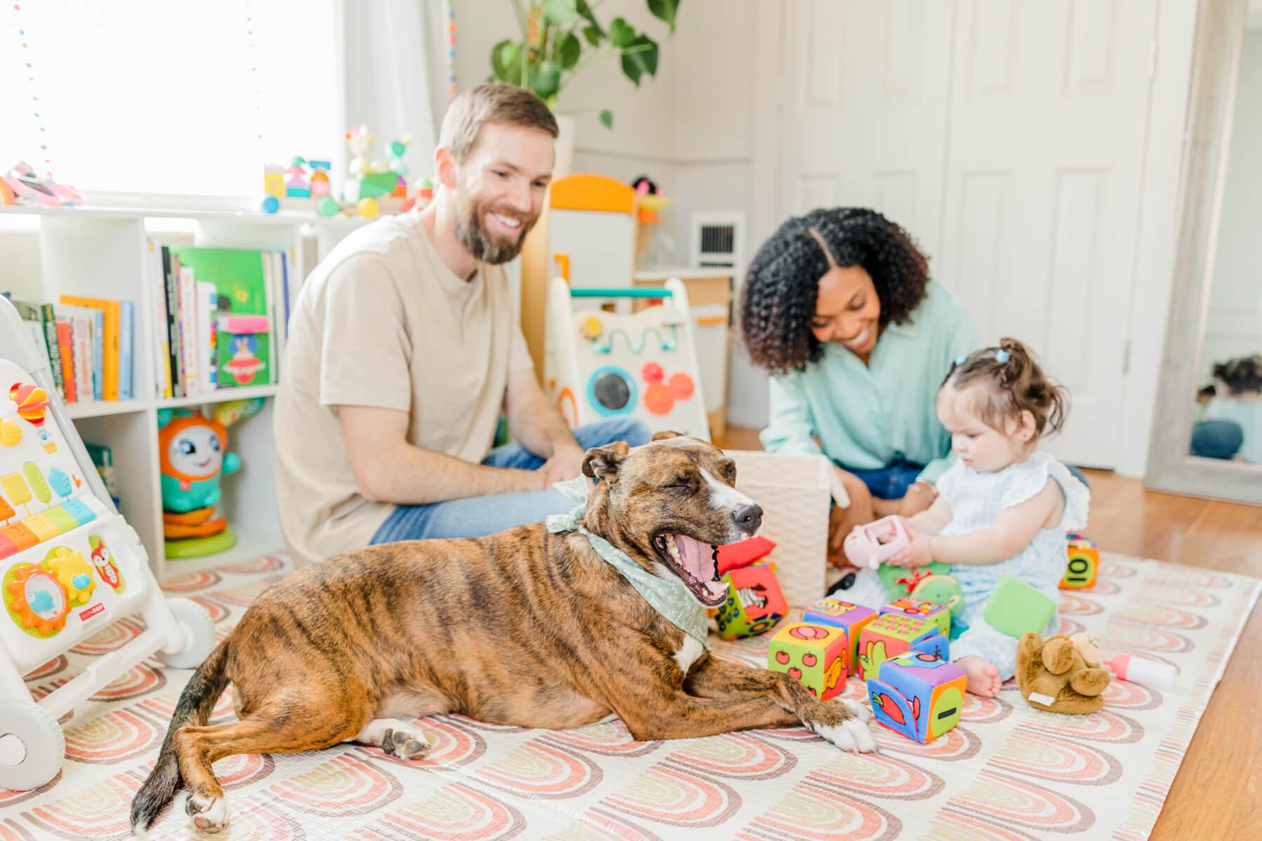 A brindle coat dog lays on the floor while his parents and infant sister play with toys in the nursery together