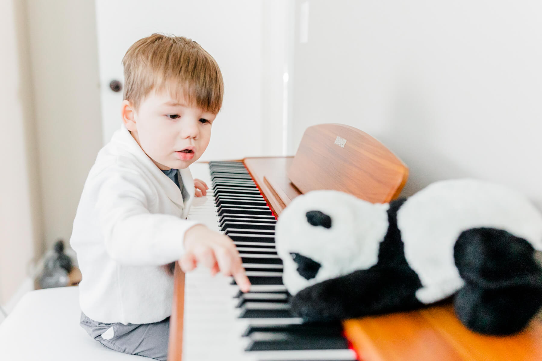 A young boy reaches for keys on a piano with his stuffed panda toy