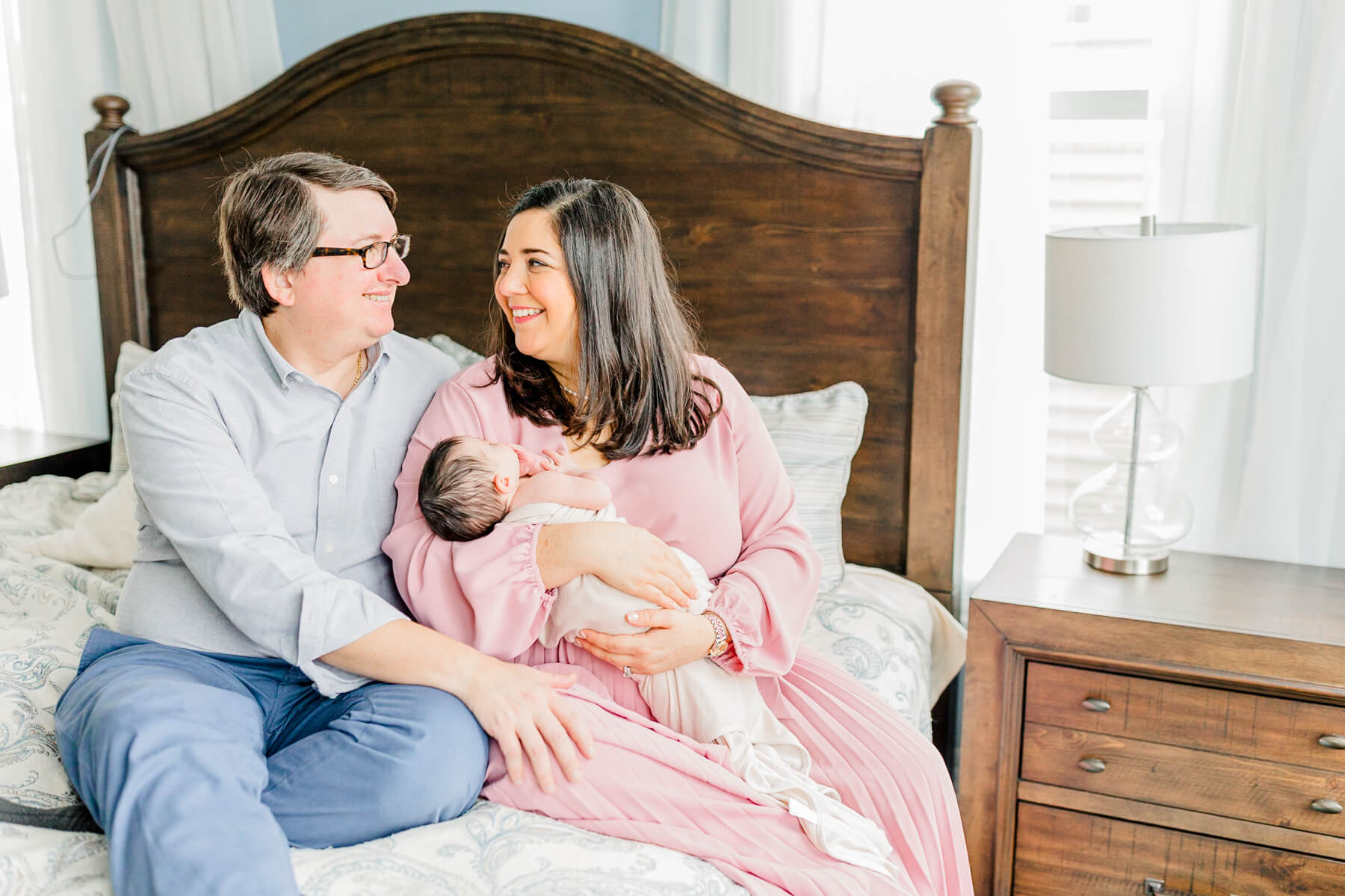 A mom and dad sit on the edge of a bed smiling at each other while mom holds their newborn baby after visiting learning express newton