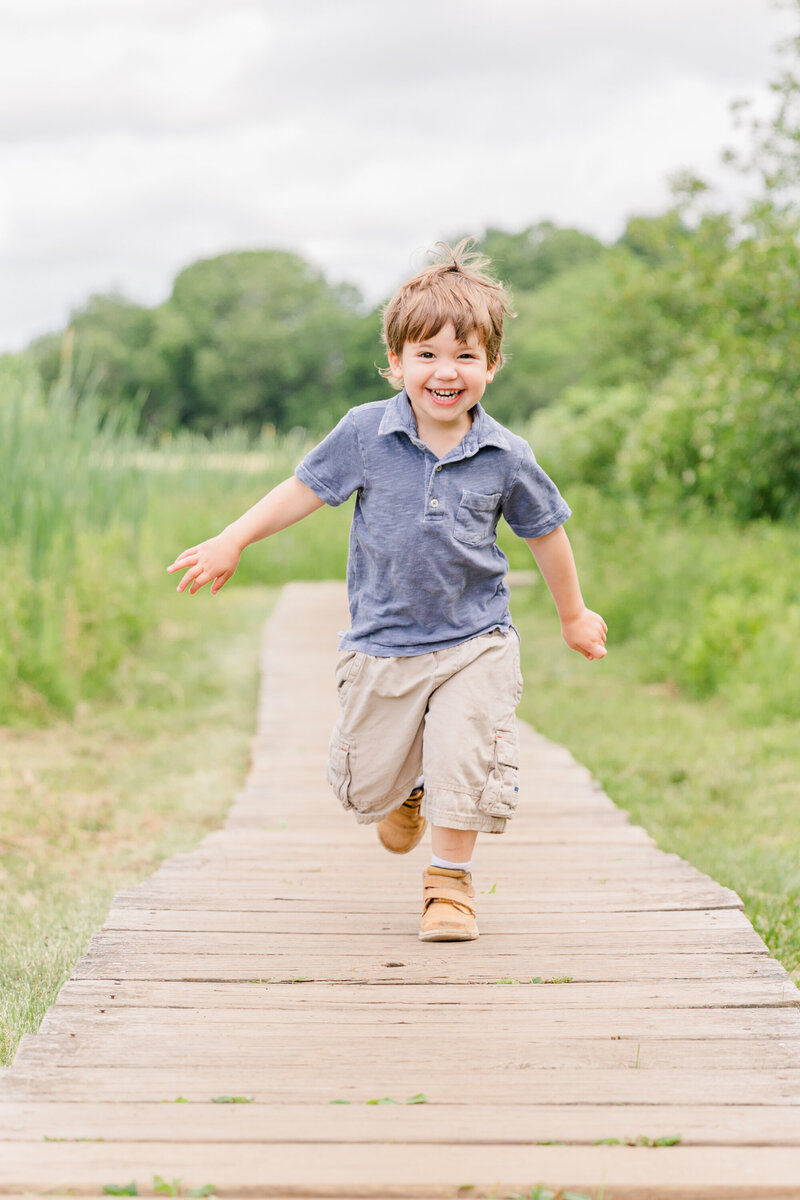 A young boy in cargo shorts runs down a boardwalk smiling in a park